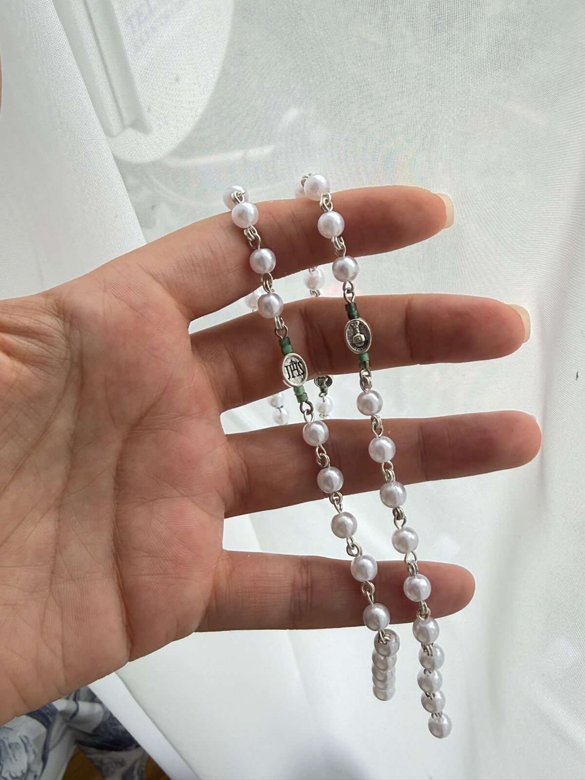 Small round white beads on fine pale-toned wire links, laid open across a palm against a sunlit curtain, with green notes at the turns and a small oval centre medal