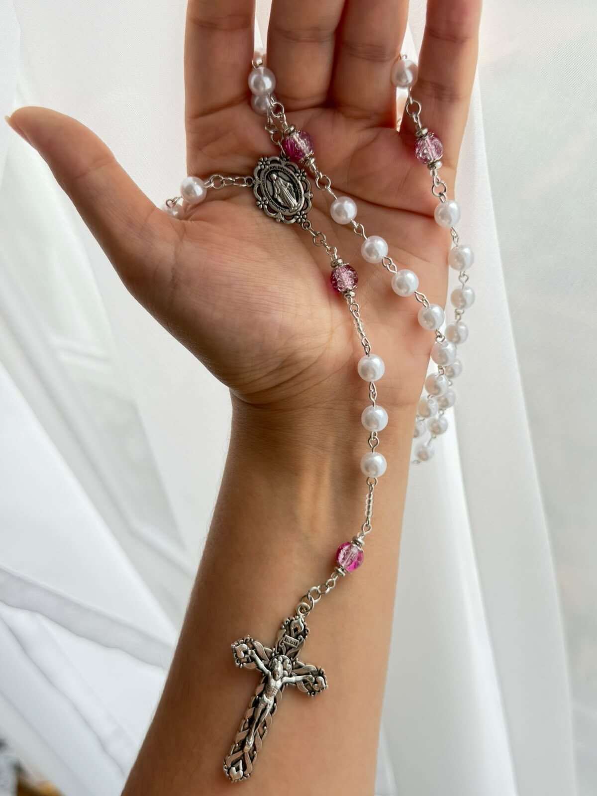 Small round white beads on fine pale-toned wire links falling across an open palm, with deep pink beads marking the turns and an ornate figured cross hanging below