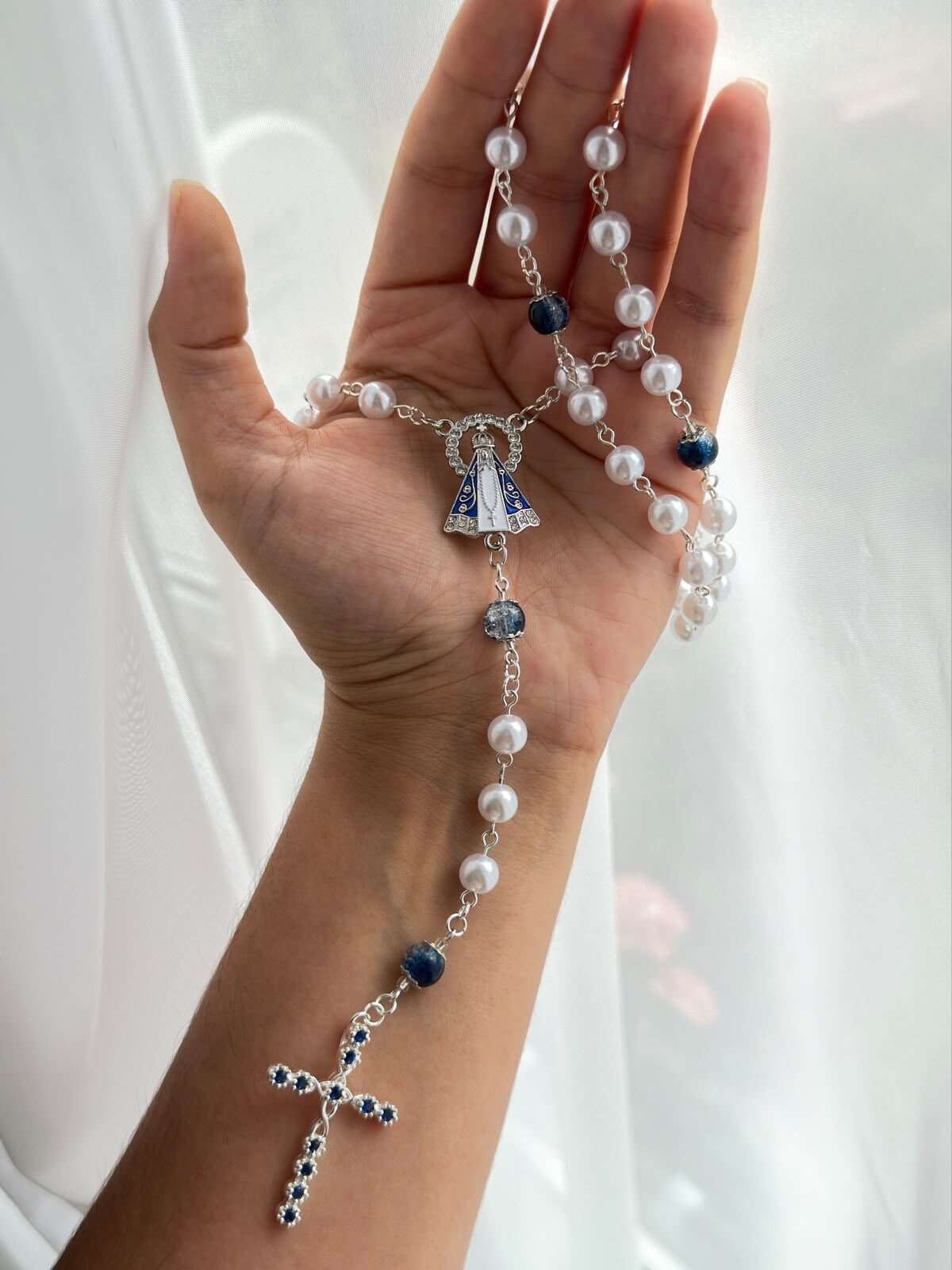Small round white beads on pale-toned wire links falling open across a palm, a deep blue bead at each turn, ending in a slender cross set with a row of small blue points