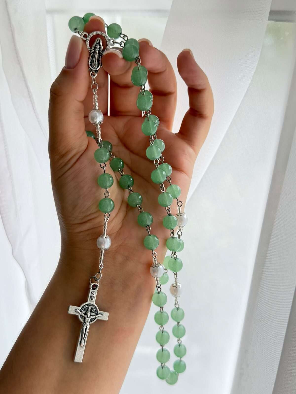 Flat green beads on fine pale-toned wire links falling down an open palm, a single white round note at each turn and a plain cross at the foot
