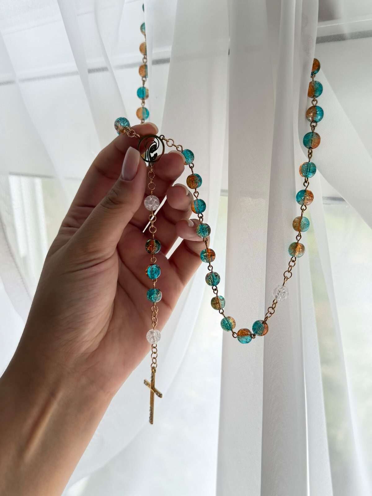 A long run of round beads shifting bead by bead from blue-green to warm amber on warm-toned links, held at an open ring centre and falling in a wide loop against a bright curtain