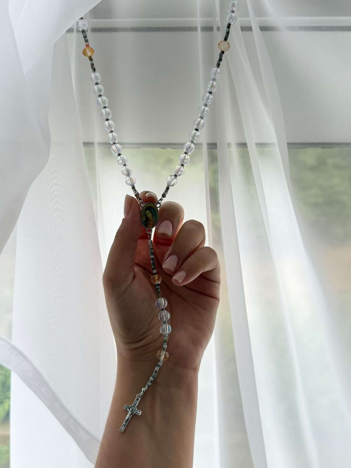A long run of nearly colourless iridescent round beads hanging at full length from a closed hand, warm amber notes recurring along it, a small painted oval centre and a dark cross at the foot