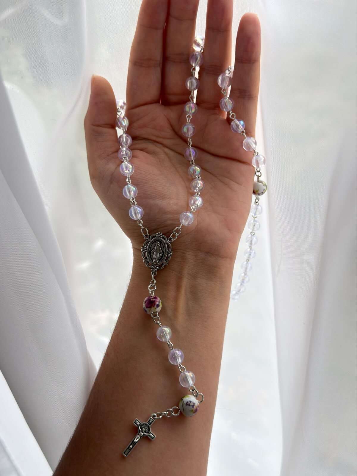 Pale iridescent round beads on fine pale-toned wire links laid down an open palm and forearm, small white beads painted with pink and violet roses at the turns, and an oval centre medal